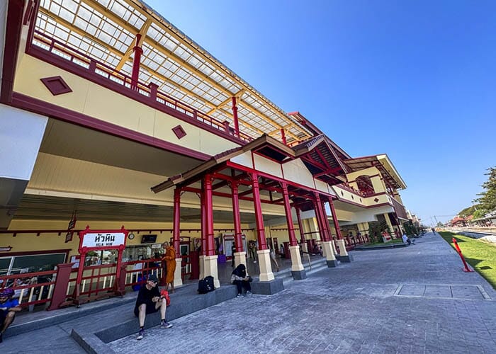 Vibrant view of Hua Hin Railway Station in Thailand, showcasing its traditional Thai architecture and a colorful SRT train approaching the platform. The station is adorned with intricate wooden designs and surrounded by lush greenery.