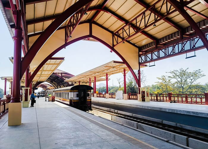 Modern Hua Hin train station with red and yellow architectural accents, a large arched roof, and a train on the tracks. A few passengers are seen on the spacious platform.